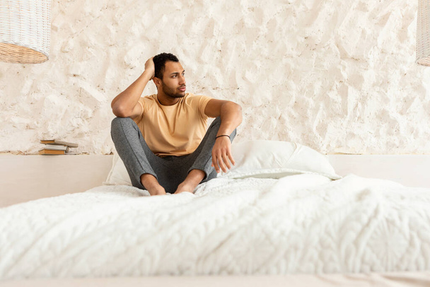 現代の寝室の屋内で無気力、欲求不満と不眠症に苦しんで、ベッドに座って脇を見ているうつ病アラビア人の男。男性の精神保健問題 - 写真・画像