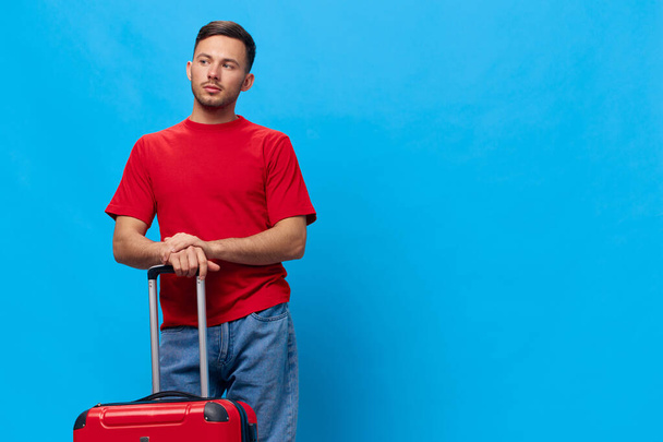 Удумливий стомлений серйозний засмучений красивий чоловік у червоній футболці чекає таксі до аеропорту з валізою, що позує ізольовано на синьому студійному фоні. Копіювати пробіл Банерний макет. концепція подорожей - Фото, зображення