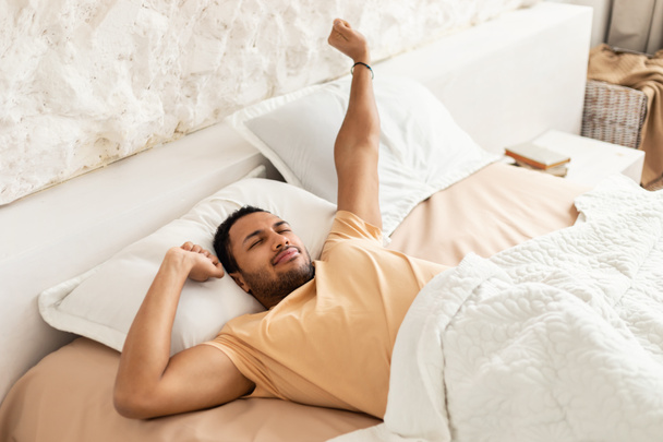 Wellslept Middle Eastern Man Awakening Yawning And Stretching Arms Lying In Cozy Bedroom Resting With Eyes Closed At Home. Good Morning. Healthy Recreation And Rest Concept - Zdjęcie, obraz