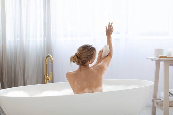 Back view of attractive blonde adult woman taking foamy bath at home, scrubbing her beautiful arms with sponge, enjoying weekend, taking care of her skin, copy space. Body care concept - Photo, image