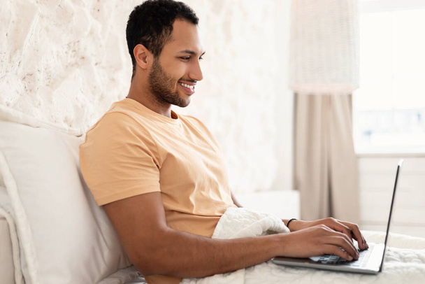 Side View Of Happy Arabic Male Freelancer Using Laptop Computer Working Online And Browsing Internet Sitting In Bedroom At Home. Freelance And Remote Job - Foto, Imagen