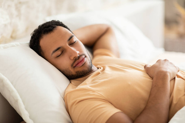 Γαλήνιος Άραβας κοιμάται κρατώντας το χέρι πίσω από το κεφάλι ξαπλωμένος με τα μάτια κλειστά στη σύγχρονη κρεβατοκάμαρα στο σπίτι. Άντρας της Μέσης Ανατολής κοιμάται στο δωμάτιό του. Έννοια υγιούς ύπνου - Φωτογραφία, εικόνα