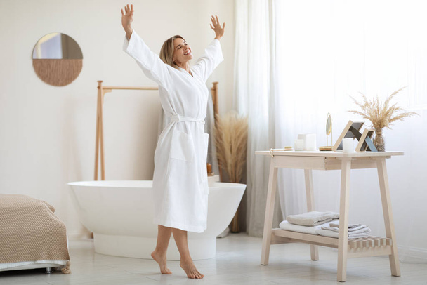 Радостная привлекательная женщина средних лет с светлыми волосами танцует с руками вверх белой ванной комнатой дома, в длинном халате, наслаждаясь выходными, собирается принять расслабляющую ванну, панораму, копировальное пространство - Фото, изображение