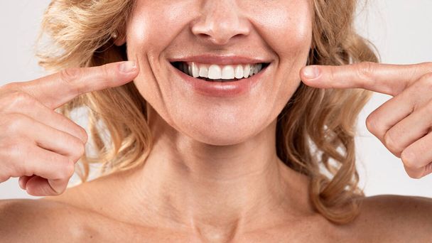 Opieka stomatologiczna. Piękna kobieta w średnim wieku wskazując na jej uśmiech z dwoma palcami, radosna dojrzała pani pokazuje białe zęby, stojąc nad jasnoszarym tle studio, przycięty strzał, zbliżenie - Zdjęcie, obraz