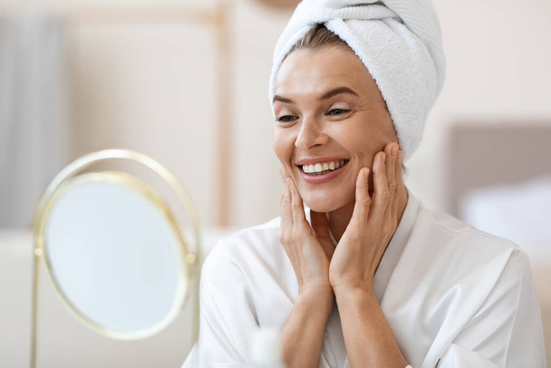 Beautiful middle aged woman in white bathrobe and with turban on her head looking at mirror, touching her face skin and smiling, using best anti-aging cosmetics, home interior, copy space - Фото, зображення