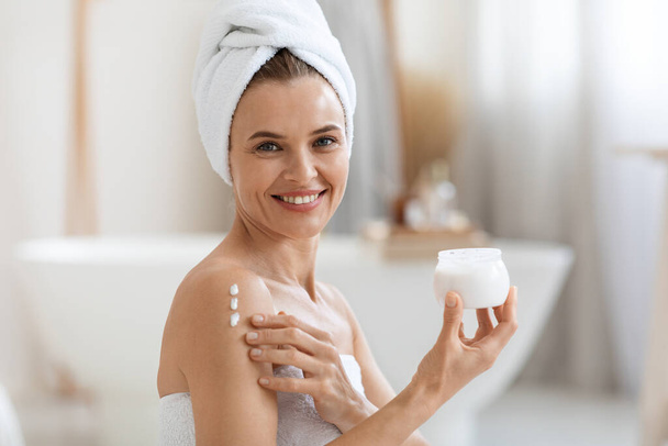 Happy good-looking adult lady wrapped in towel using body lotion after shower, beautiful middle aged woman applying moisturizing or nourishing cream on her shoulder, copy space - Fotó, kép