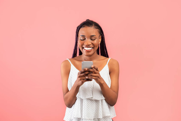 Ευτυχισμένη νεαρή μαύρη γυναίκα που χρησιμοποιεί smartphone, συνομιλεί στο διαδίκτυο, βλέπει βίντεο σε ροζ φόντο στούντιο. Lovely African American Lady surfing web, αποστολή sms, απόσπαση στα μέσα κοινωνικής δικτύωσης - Φωτογραφία, εικόνα