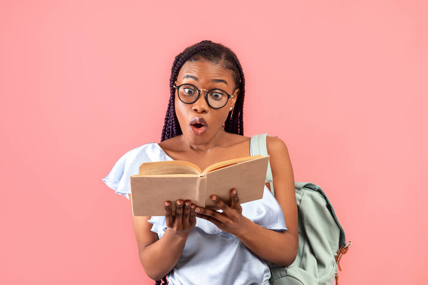 Потрясенная афроамериканка с рюкзаком, смотрящим в открытый учебник на розовом фоне студии. Удивлённая чернокожая женщина чувствует себя подавленной университетским или университетским образованием - Фото, изображение