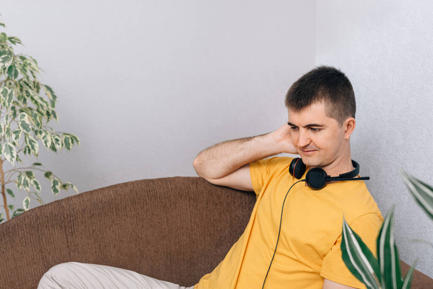 Νεαρός χαμογελαστός άντρας με κίτρινο μπλουζάκι που φοράει ακουστικά στο σπίτι. Το αγόρι ακούει μουσική ή διαλέξεις. Πράσινα φυτά στο δωμάτιο. Online εκπαίδευση και η έννοια της οικιακής κηπουρικής. - Φωτογραφία, εικόνα