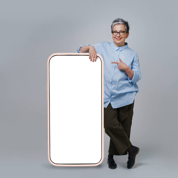Mature grey hair woman pointing finger on screen of giant, huge smartphone with white, blank screen standing leaned on wearing casual isolated on white background. Free space mock up. - Valokuva, kuva