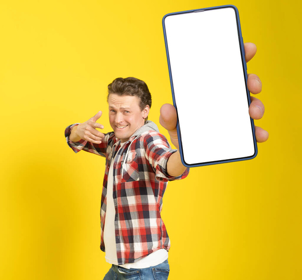 Happy to win handsome man pointing at smartphone with white empty screen, wearing red plaid shirt and jeans cellphone display mock up isolated on yellow background. Mobile app advertisement.  - 写真・画像