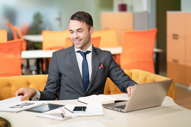 Young business man in suit, sitting at his desk, looking at documents. With laptop on the table, documents, papers, office supplies, a smartphone and tablet pc. Business concept.  - Foto, immagini