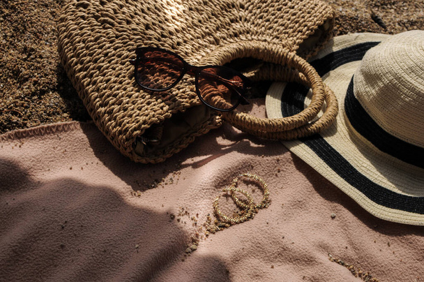 Top view of beach accessories: straw hat, summer bag, sunglasses, and earrings on bed cover on sand beach on a summer day  - Foto, Bild