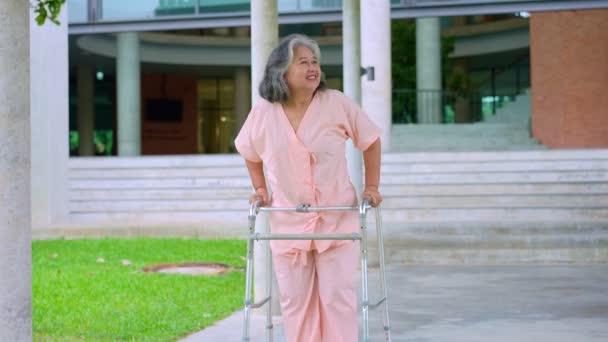A boldog idős ázsiai nő járókereket használ, és besétál a kórházba, miután felépült egy balesetből. A boldog nyugdíjazás fogalma Gondozás a gondozótól, a megtakarításoktól és a nyugdíjas egészségbiztosítástól - Felvétel, videó