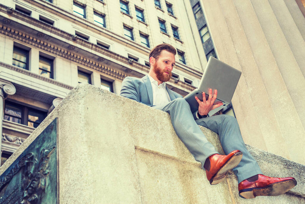 American Businessman with beard, mustache traveling, working in New York, wearing cadet blue suit,  white undershirt, brown leather shoes, sitting on street, reading, working on laptop computer. - Foto, Imagen
