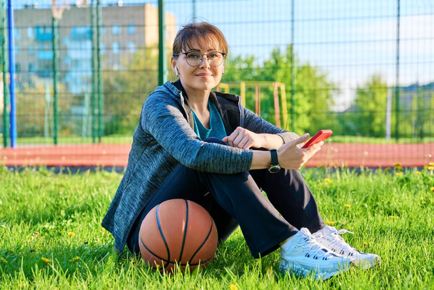 Active mature woman with basketball ball and smartphone, outdoor basketball court background. Middle-aged female in sportswear headphones sitting on grass. Sporty healthy lifestyle, leisure activity - 写真・画像