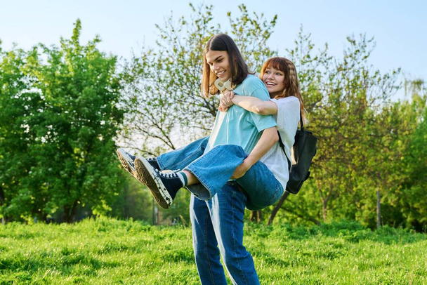 Happy laughing youth, having fun couple of teenagers outdoor. Beautiful young girl friends on boyfriends back enjoying together, green lawn sky background. Summer, love vacation, young people concept - 写真・画像