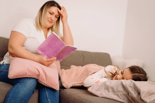 Mother reading to her child daughter book of pink color sitting on the sofa while little daughter sleeping. . High quality photo - Zdjęcie, obraz
