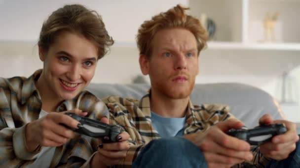 Vidám feleség kezében gamepad harcol zavaros vörös hajú férj otthon közelkép. Izgatott nő élvezi virtuális verseny frusztrált vörös férfi. Barátok segítségével joysticks játszani videojáték hétvégén - Felvétel, videó
