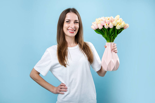 Усміхнена молода жінка тримає вазу з тюльпанами на синьому фоні
. - Фото, зображення