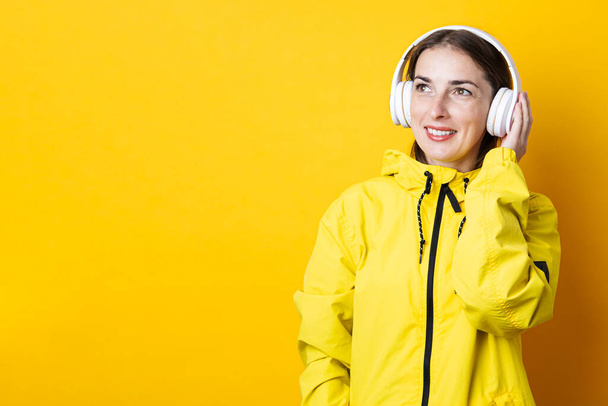 Smiling young woman in headphones in a yellow jacket looks up on a yellow background - Фото, зображення