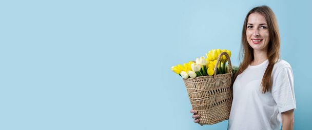 Uśmiechnięta młoda kobieta trzymająca żółte tulipany w koszyku na niebieskim tle. Sztandar. - Zdjęcie, obraz