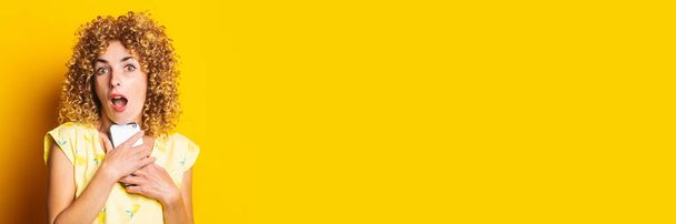 emotional beautiful curly young woman with a phone on a yellow background. Banner - Φωτογραφία, εικόνα