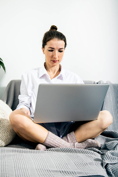 Jonge vrouw in wit shirt met laptop zittend op de bank in de kamer. - Foto, afbeelding