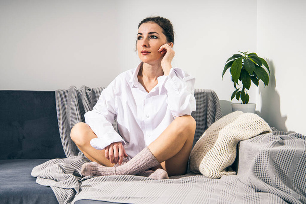 Jeune femme réfléchie en chemise blanche en chaussettes tricotées assis sur le canapé dans la chambre. - Photo, image