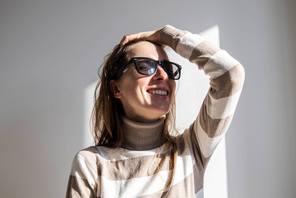 Smiling young woman in sunglasses fixing her hair against a white wall  - Photo, Image