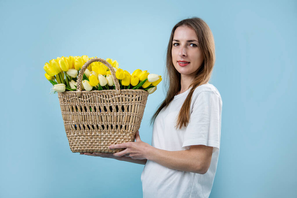 Усміхнена молода жінка тримає жовті тюльпани в кошику на синьому фоні
. - Фото, зображення