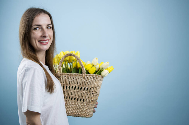Lächelnde junge Frau mit gelben Tulpen im Korb auf blauem Hintergrund. - Foto, Bild