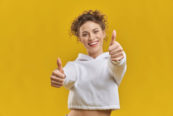 Young curly lady doing positive gesture with hands, approving indoors. Portrait view of joyful female feeling positive with both thumbs up, isolated on yellow studio background. Concept of gesturing.  - Zdjęcie, obraz