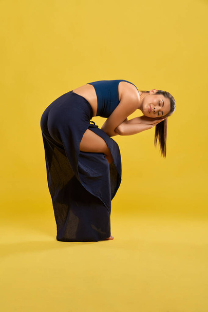 Flexible woman twisting body and arms in asana of yoga, while training indoor. Side view of slim female with closed eyes, practicing yoga, meditating, isolated on orange background. Concept of yoga.  - Foto, immagini