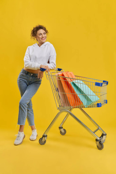 Cute girl in casual clothes, leaning on cart with purchases in studio. Front view of brunette lady with crossed legs, holding trolley, looking away, isolated on orange background. Concept of shopping. - Foto, afbeelding