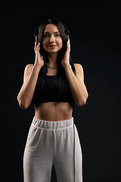 Happy girl in crop top, looking at camera, while listening to music inside. Portrait view of slim female holding headphones, enjoying sound, isolated on black studio background. Concept of music fan. - Photo, image