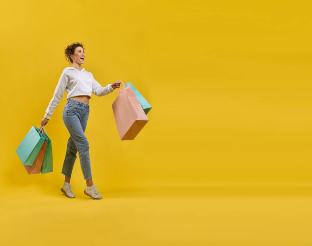Delighted female shopper with paper bags walking alone after shopping in studio. Side view of trendy woman carrying shopping bags, looking ahead, isolated on orange background. Concept of shopping. - Фото, зображення