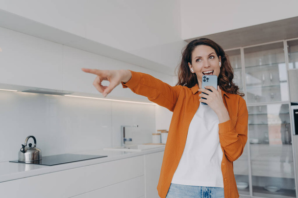 Carefree spanish woman in earphones is singing with cellphone as with microphone. Application for music listening online. Playful passionate girl is singing at home and points to you. - Foto, immagini