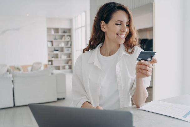Szczęśliwa Europejka trzyma kartę kredytową i kupuje przez internet. Zakup przez internet na laptopie. Pozytywna kobieta korzystająca z bankowości internetowej. Koncepcja handlu elektronicznego i transakcji finansowych. - Zdjęcie, obraz