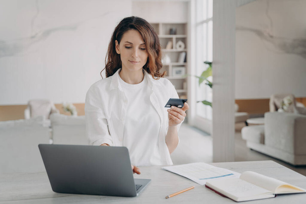 Young pretty woman in white is going to buy through internet. European girl is holding credit card in front of laptop. Booking or purchasing online. E-commerce and financial transaction concept. - Foto, Imagen