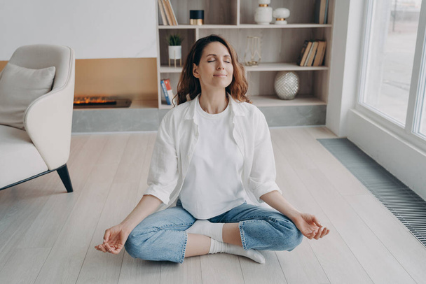 Girl is sitting in lotus pose with her eyes closed. Happy european woman is practicing yoga and meditation on floor at home. Morning gymnastics and zen. Stress relief, wellbeing and peace concept. - Photo, Image
