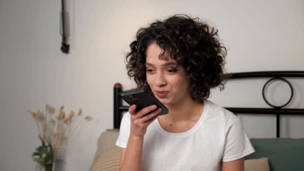 Smiling hispanic curly woman student uses smartphone dictating an audio message to friend. Positive beautiful female holding mobile phone in hand talking, sitting on bed in bedroom at home - Imágenes, Vídeo