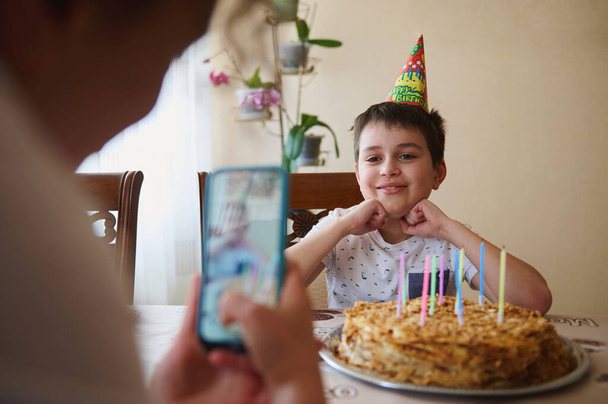 Allegro adorabile bel ragazzo caucasico adolescente siede a tavola con torta di compleanno con candele, sorride mentre posa alla sua mamma sfocata scattandogli foto sullo smartphone in primo piano. Festa di compleanno - Foto, immagini