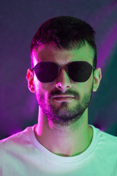 Portrait of a young Caucasian man with beard wearing sunglasses and looking towards the camera illuminated with green and magenta colored lights. - Fotografie, Obrázek
