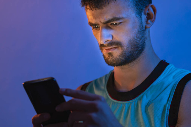 Portrait of a young Caucasian man with beard using his cell phone. It is illuminated with blue and orange colored lights. - Foto, Bild