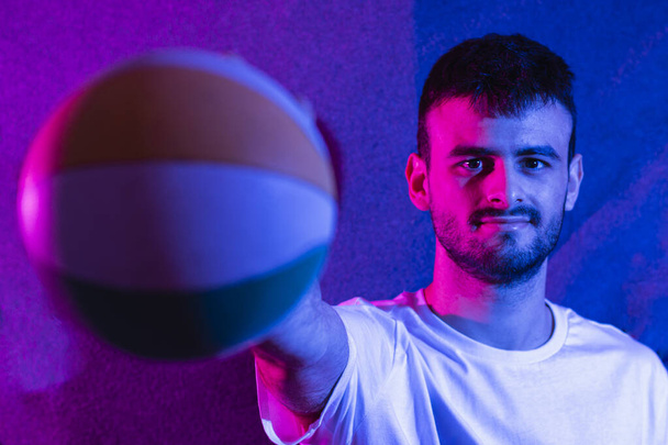 Een jonge blanke bebaarde man met uitgestrekte arm met een kleine basketbal van verschillende kleuren. De bal is uit focus op de voorgrond en de persoon is in focus. Er zijn gekleurde lichten.. - Foto, afbeelding