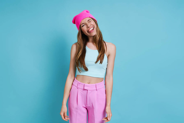 Playful young woman in pink hat sticking out tongue while standing against blue background - 写真・画像