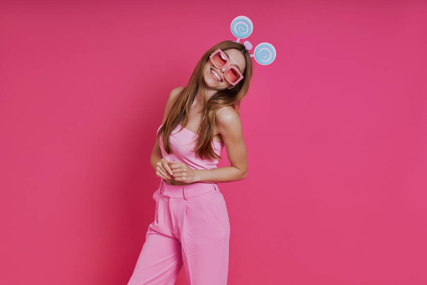 Funky young woman in party headwear smiling while standing against pink background - Foto, afbeelding