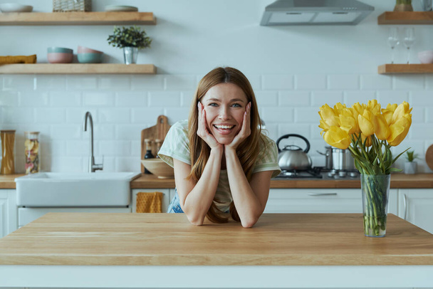 Happy young woman leaning at the kitchen desk and smiling - Valokuva, kuva
