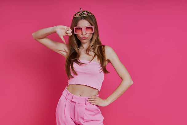 Unhappy young woman in funky crown showing her thumb down while standing against pink background - Foto, Imagen